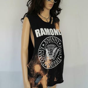 May include: Black sleeveless top with the band name "Ramones" in white. The shirt has a distressed, bleached effect and a circular graphic with the words "Tommy, Johnny, Dee Dee" around a crest. A punk rock style.