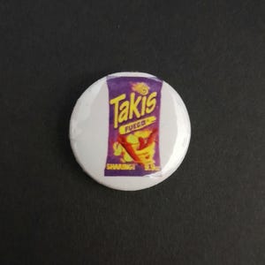 May include: A round white button badge featuring a purple and yellow graphic of a Takis Fuego bag. The bag design includes red chilli peppers and the words "Takis Fuego". The badge has a shiny surface.