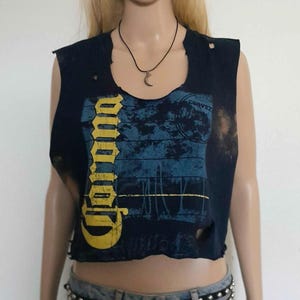 May include: A blue sleeveless crop top with a distressed look. The top features a yellow graphic of the word "Corona" and a blue and white graphic with the word "Cerveza".