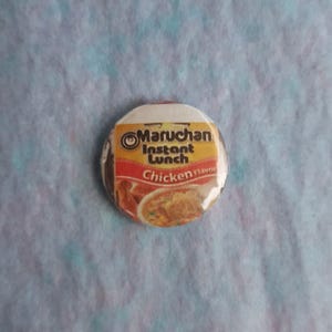 May include: A circular button displaying the Maruchan Instant Lunch Chicken Flavour design. The button has a white top and a red and yellow label with the brand name and product description. The image of the chicken flavour soup is visible.