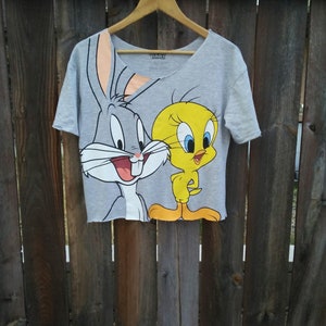 Może przedstawiać: Szara koszulka crop top z Królikiem Bugsem i Tweety Bird z Looney Tunes. Koszulka ma surowe obszycie i dekolt w kształcie litery V. Postacie z kreskówek są w kolorze na szarym tle.