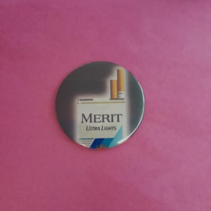 May include: A circular badge displaying a Merit Ultra Lights cigarette packet. The badge has a silver-grey background. The packet is white with the brand name and 'Ultra Lights' text. The background is a solid pink colour.