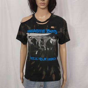 May include: Black tie-dye t-shirt with a distressed neckline and shoulder cut-out. The shirt features the band name "BEASTIE BOYS" in blue, along with a black and white image and the text "CHECK YOUR HEAD" in blue.