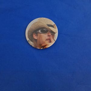 May include: A circular badge showing a person wearing a straw hat and sunglasses, with a finger to their lips. The badge has a light background and is set against a blue background.