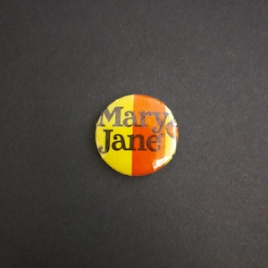 May include: A circular badge featuring the words "Mary Jane" in black. The badge is split into yellow and orange halves. The badge is set against a black background.