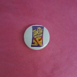 May include: A white, round button featuring a graphic of a Takis Fuego bag. The bag is purple with the word "Takis" in yellow and red chili peppers. The button is set against a pink background.