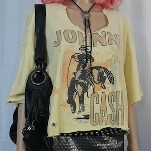 Könnte beinhalten: Gelbes, gecropptes T-Shirt mit einem Used-Design, das eine Grafik eines Cowboys auf einem Pferd zeigt, mit dem Text "JOHNNY CASH". Das Shirt wird mit einem schwarzen Rock und einer schwarzen Umhängetasche kombiniert.