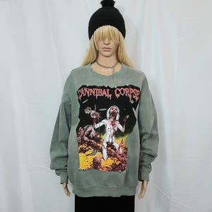 Puede incluir: Una sudadera verde descolorida con el texto "CANNIBAL CORPSE" y una ilustración gráfica. El gráfico muestra una escena perturbadora con figuras en blanco, rojo y marrón. La modelo lleva un gorro negro.
