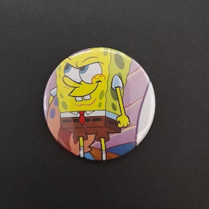 Puede incluir: Un botón redondo con una imagen de dibujos animados de Bob Esponja con una expresión traviesa. El botón tiene un fondo amarillo con un atuendo marrón y rojo. El fondo es una mezcla de rosa, morado y blanco.