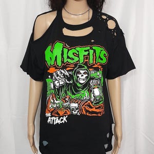May include: Black distressed t-shirt featuring the band name "Misfits" in green block letters above a graphic of a grim reaper holding a lantern. The words "The Attack" are below the graphic.