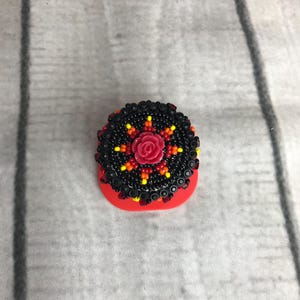 May include: A small, red, beaded object with a floral design. The top features a circular pattern of black beads with yellow, orange, and red accents, surrounding a central red rose. The base is a solid, bright red color.
