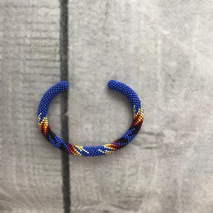 May include: A blue beaded cuff bracelet with a colorful pattern of yellow, red, and black. The bracelet is open-ended and appears to be handmade, showcasing a detailed beadwork design. The bracelet is set against a neutral, textured background.