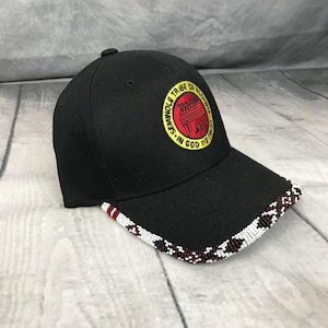 May include: A black baseball cap featuring a circular patch with a red center and yellow border displaying the text Seminole Tribe of Florida In God We Trust. The brim is decorated with a beaded pattern in white, black, and dark red colors.
