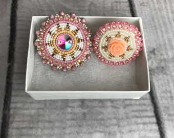 May include: Two beaded circular brooches in a white box. One brooch features a central iridescent gem surrounded by white, gold, and pink beads. The other has a peach-colored rose in the center, surrounded by white beads and a pink beaded border.