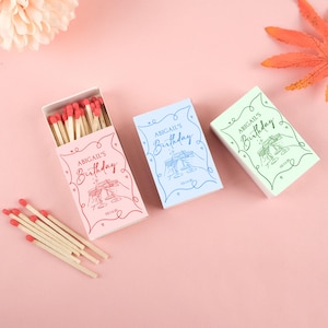 Könnte beinhalten: Drei Streichholzschachteln in Rosa, Blau und Grün, jeweils mit dem Text "ABIGAIL'S Birthday" in Schreibschrift. Die rosa Schachtel ist geöffnet und zeigt rotköpfige Streichhölzer. Die Schachteln haben einen dekorativen Rahmen und eine Champagnerglas-Illustration.