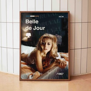 May include: A framed poster for the film "Belle de Jour", featuring a photograph of a woman. The poster is in a dark brown frame, with the year "/1967" at the bottom. The background is a tiled wall.