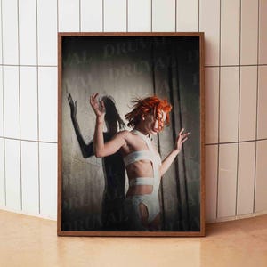 Könnte beinhalten: Gerahmter Kunstdruck mit einer Person mit roten Haaren in einem weißen Outfit, die Arme erhoben. Der Schatten der Person wird auf die dahinterliegende Wand geworfen. Der Druck befindet sich in einem dunkelbraunen Rahmen.