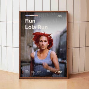 Könnte beinhalten: Gerahmtes Filmplakat für "Lola rennt", das eine Frau mit leuchtend roten Haaren beim Laufen zeigt. Das Poster zeigt den Filmtitel in Weiß, ein blaues Tanktop und das Jahr 1998.