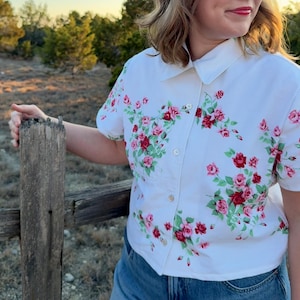 May include: White short-sleeved shirt with a floral pattern of pink and red roses and green leaves. The shirt has a button-down front and a collar. The shirt is paired with blue denim jeans.