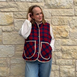 May include: A red and navy houndstooth pattern vest with cream-colored trim and pockets. The vest is worn over a white blouse and paired with blue jeans. The vest has a button-up front and a high collar.
