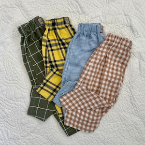 May include: Four pairs of patterned pajama pants in various colors. One pair is olive green with a white grid pattern, another is yellow with a black plaid, a third is light blue with a gingham check, and the last is brown and white gingham.