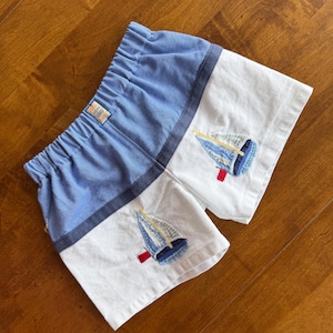 May include: Blue and white shorts with an elastic waistband. The shorts feature embroidered sailboat designs on the white panels. The shorts are made of a soft fabric and are likely for children.