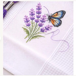 Puede incluir: Tela blanca bordada a punto de cruz con un dise&ntilde;o de lavanda y mariposa. La lavanda es morada con hojas verdes, y la mariposa tiene alas azules, naranjas y negras. Se ven una aguja e hilo morado.