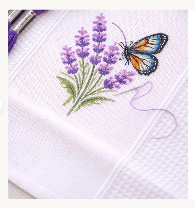 Puede incluir: Tela blanca bordada a punto de cruz con un dise&ntilde;o de lavanda y mariposa. La lavanda es morada con hojas verdes, y la mariposa tiene alas azules, naranjas y negras. Se ven una aguja e hilo morado.