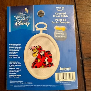 May include: A Disney Tigger counted cross stitch kit. The kit includes a white oval frame with a cross-stitched image of Tigger in red and yellow. The finished size is 2.25" x 2.75". The kit includes fabric, floss, needle, and instructions.