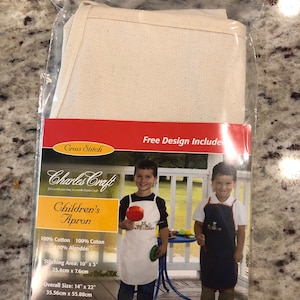 May include: A white cotton children's apron with a 10 inch by 3 inch stitching area. The apron is 14 count and comes with a free design. The packaging shows two children wearing aprons, one white and one blue, with a red and blue toy truck design.