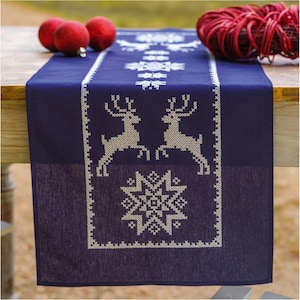 May include: A navy blue table runner with a white cross-stitch design featuring reindeer and snowflakes. The runner is rectangular and has a decorative border. Red Christmas ornaments and a red ball of twine are in the background.