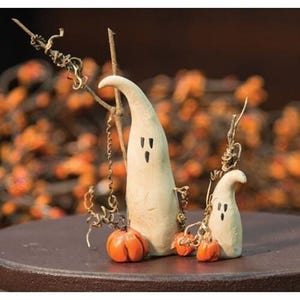 May include: A pair of off-white ghost figurines with black eyes and mouths, accompanied by small orange pumpkins. The ghosts are decorated with twigs and vines, creating a whimsical Halloween or fall theme.