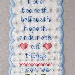 May include: White cross-stitch bookmark with blue scalloped edges. It features a floral design with blue flowers, green leaves, and pink hearts. The text reads "Love beareth, believeth, hopeth, endureth all things 1 COR 13:7".