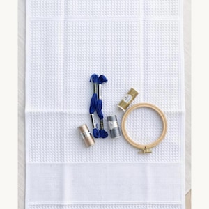 Puede incluir: Una tela blanca de tejido de gofre con un dise&ntilde;o de punto de cruz, acompa&ntilde;ada de hilos de bordar azules, plateados y dorados, y un bastidor de bordado de madera. Los suministros de manualidades est&aacute;n dispuestos sobre la tela blanca, listos para un proyecto de costura.