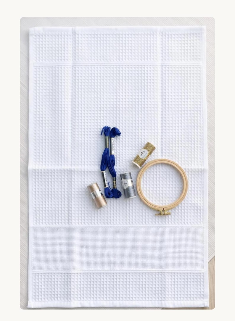 Puede incluir: Una tela blanca de tejido de gofre con un dise&ntilde;o de punto de cruz, acompa&ntilde;ada de hilos de bordar azules, plateados y dorados, y un bastidor de bordado de madera. Los suministros de manualidades est&aacute;n dispuestos sobre la tela blanca, listos para un proyecto de costura.