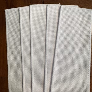 May include: Five rectangular pieces of white Aida cloth arranged in a fan shape. Each piece features a distinct, even grid pattern of small holes designed for cross-stitch embroidery, with finished, serged edges along the vertical sides.
