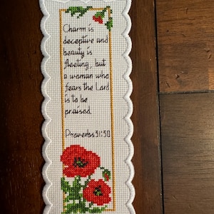 May include: White cross-stitch bookmark with a scalloped edge. The bookmark features the text "Charm is deceptive and beauty is fleeting, but a woman who fears the Lord is to be praised" and "Proverbs 31:30". The bottom of the bookmark has a cross-stitched image of red poppies.