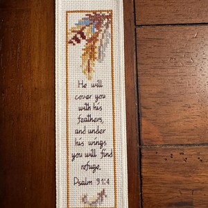 May include: A rectangular white fabric bookmark featuring a cross-stitched design of a feather at the top and a small bird at the bottom. The text reads: He will cover you with his feathers, and under his wings you will find refuge. Psalm 91:4.