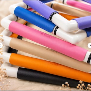May include: A stack of square embroidery frames featuring white plastic corner joints and straight segments covered in colorful foam grips. The grips are colored purple, blue, pink, tan, orange, and black, each embossed with the Nurge brand name.