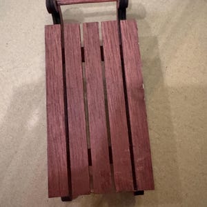 May include: A miniature wooden sled with a reddish-brown deck and a handle. The sled has black runners and a simple design, suitable for decorative purposes or small toys.
