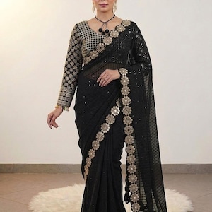 May include: A black saree with gold floral embroidery along the borders and sleeves. The saree features a sheer, patterned fabric and a matching blouse with intricate gold detailing. The overall design is elegant and formal.
