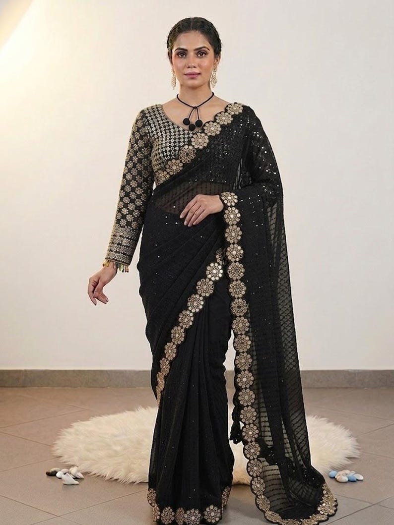 May include: A black saree with gold floral embroidery along the borders and sleeves. The saree features a sheer, patterned fabric and a matching blouse with intricate gold detailing. The overall design is elegant and formal.