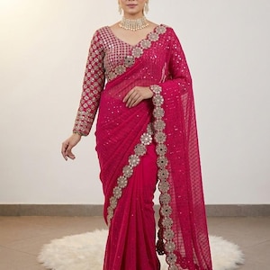 May include: A magenta sari with intricate gold embroidery and embellishments. The long-sleeved blouse features a matching design. The sari's border is detailed with a scalloped edge and decorative accents. The outfit is worn with a matching necklace and earrings.