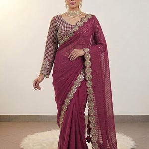 May include: A burgundy saree with intricate gold embroidery and embellishments. The saree features a detailed border and a matching long-sleeved blouse. The fabric drapes elegantly, showcasing the rich color and ornate design. The saree is a traditional garment.