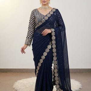 May include: A navy blue saree with intricate gold embroidery and sequin embellishments. The long-sleeved blouse features a silver and gold pattern. The saree is draped elegantly, showcasing the detailed design and texture of the fabric.