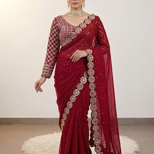May include: A deep red saree with intricate gold embroidery and embellishments. The long-sleeved blouse features a matching design. The saree's border is adorned with circular gold accents. The overall design is elegant and formal.
