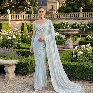 May include: A pale blue saree with detailed white embroidery and a flowing train. The saree has long sleeves and a subtle neckline. The woman is in a garden with a stone bench and fountain in the background.