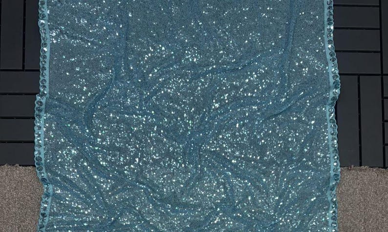 Puede incluir: Una tela azul claro brillante, densamente cubierta de lentejuelas. La tela est&aacute; drapeada, creando pliegues y sombras suaves. Un borde decorativo con peque&ntilde;as lentejuelas a juego adorna el borde. El efecto general es de elegancia y brillo.