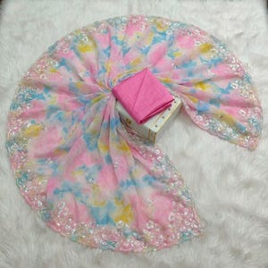 May include: A vibrant, tie-dye patterned fabric in shades of pink, blue, and yellow, with a delicate, sequined border. A solid pink fabric is neatly folded and placed on a small, white box. The fabric is arranged on a white, fluffy surface.