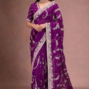 May include: A vibrant purple saree with intricate white floral embroidery. The saree features a detailed border and matching short-sleeved blouse. The fabric drapes elegantly, showcasing the floral design.